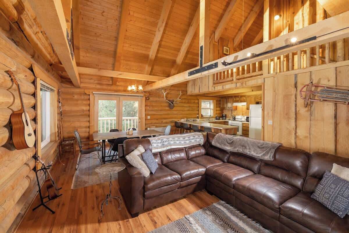A log cabin interior with a leather sofa, wooden walls, a dining area, and a kitchen.