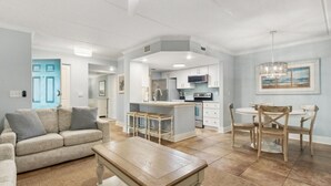 Un vaste espace de vie comprenant un canapé, une table basse et une table à manger avec des chaises. On aperçoit une cuisine à l’arrière-plan, avec une porte bleue et un luminaire suspendu.