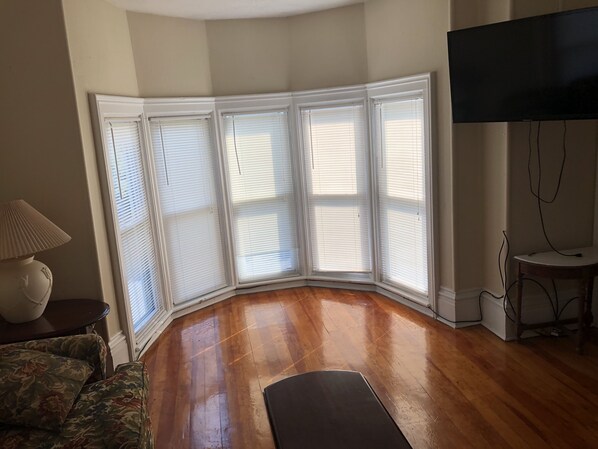 A room with a corner bay window, wooden flooring, a flat-screen TV, and a lamp. - 1 bedroom/1 bath (Oneonta)