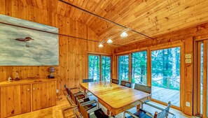 Un interior de madera con una gran mesa de comedor, sillas metálicas y ventanas amplias que dan a un bosque.