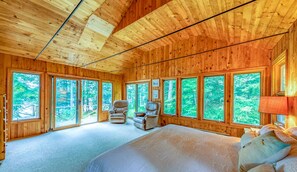 Un dormitorio de cabaña de madera con una cama grande, dos sillones y varias ventanas que dan a vistas del bosque.