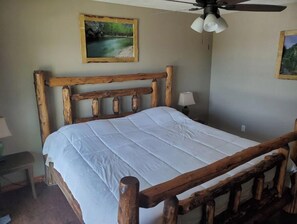 Una cama con estructura de madera y colchón blanco en una habitación con ventilador de techo y dos cuadros de paisajes en las paredes.