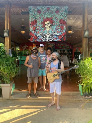 A group of people standing in a covered outdoor area with a large colorful tapestry and potted plants.