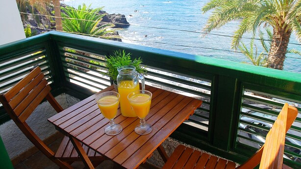 Una mesa de madera con dos vasos de jugo de naranja y una jarra, sobre un balcón con vista al océano y palmeras.