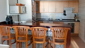 Cocina compacta con armarios de madera, microondas, cafetera y comedor con sillas.