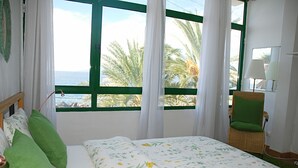 Un dormitorio con una cama, almohadas verdes, una silla de mimbre, una lámpara y una ventana con vistas a palmeras y al mar.