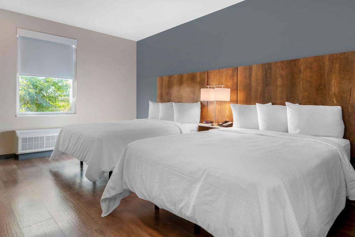 Habitación de hotel con dos camas, cabecero de madera, ventana con persianas y mesita de noche con lámpara.