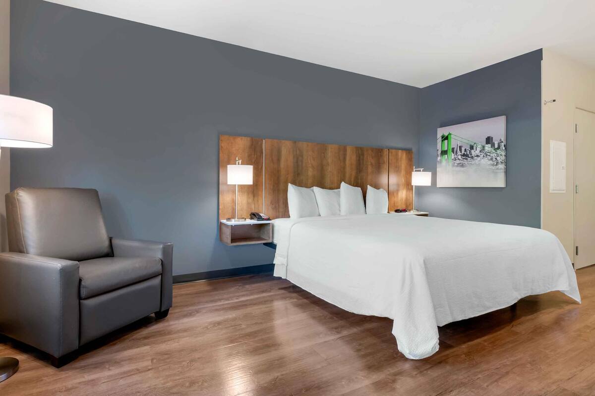 Una habitación de hotel con una cama grande, un sillón gris, una mesita de noche con una lámpara y una estantería en la pared.