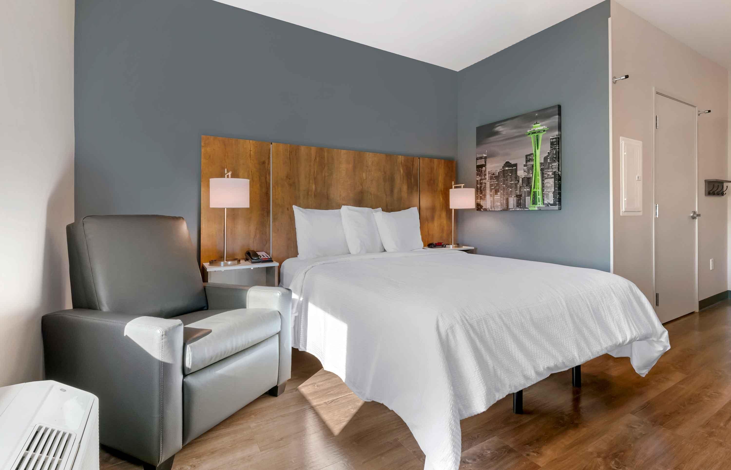 Una habitació d'hotel amb llit, cadira, tauleta de nit, làmpada i un quadre d'un paisatge urbà.