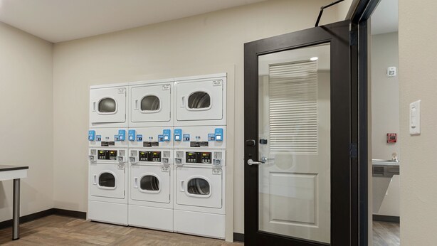 Une buanderie équipée de plusieurs machines à laver et de plusieurs sèche-linge, une porte dotée d’un système d’alarme incendie et une petite table.