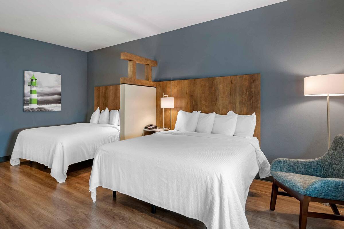 Habitación de hotel con dos camas, cabecero de madera, mesita de noche, lámpara y una silla.