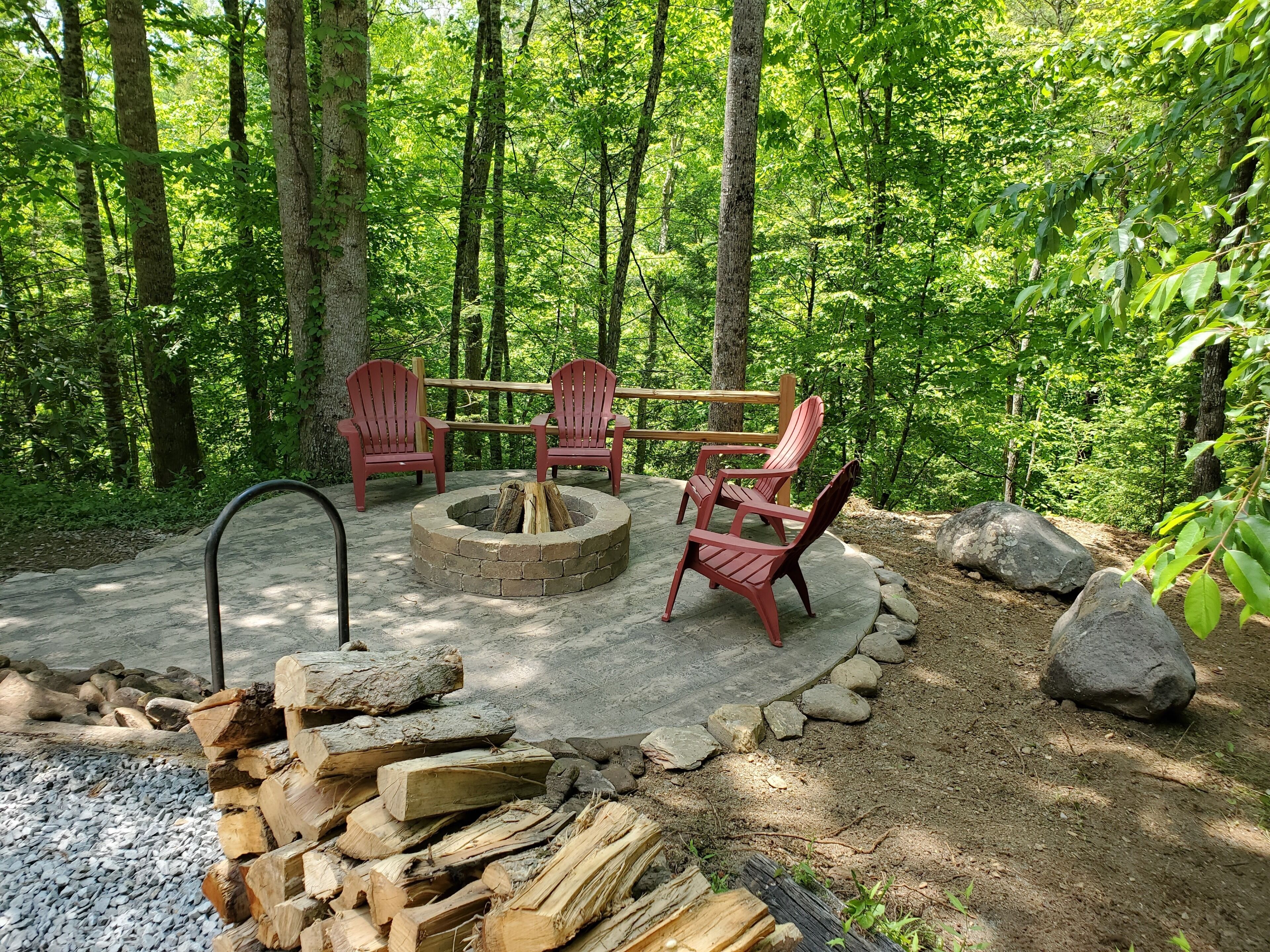 A circular fire pit with chairs around it, set in a forested area with a stone path and wooden logs nearby.