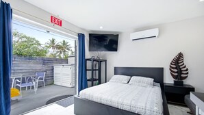 A modern hotel room with a bed, a TV, an air conditioner, a desk, and a view of the outdoors. - Total Oasis - 8BR Resort Compound, Pool & Tiki Bar, Sleeps 30, Near Cruise Port (Hollywood)