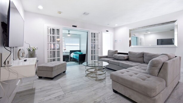 A modern living room with a sofa, ottoman, and glass coffee table. - Total Oasis - 8BR Resort Compound, Pool & Tiki Bar, Sleeps 30, Near Cruise Port (Hollywood)