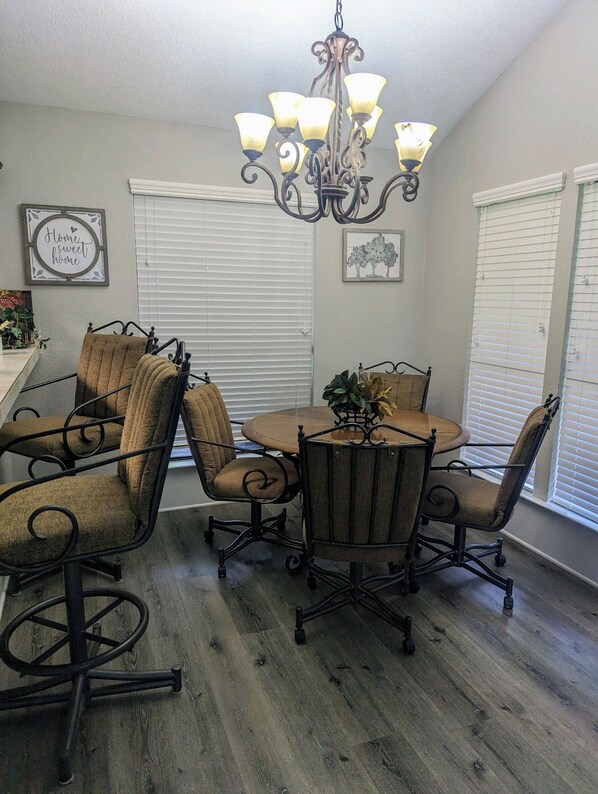 A dining area with a round table and four chairs, a chandelier, and a window with blinds.