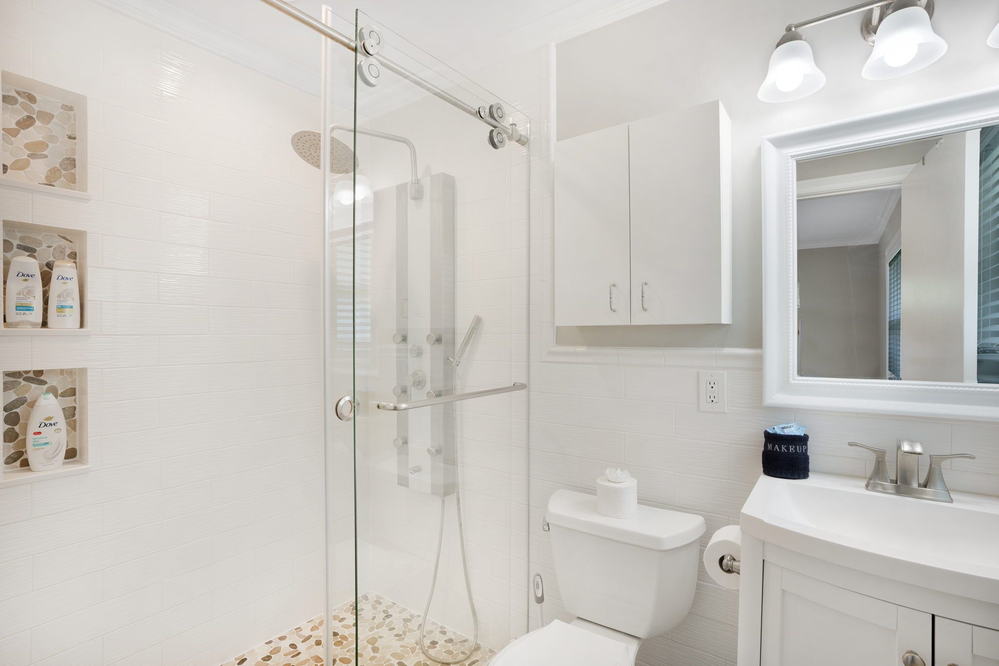 A modern bathroom with a glass-enclosed shower, white tiles, a vanity with a mirror, and Dove toiletries on shelves.