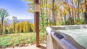 Un bain à remous sous une tonnelle en bois, agrémenté d’un panneau de sécurité, entouré d’arbres aux couleurs automnales et offrant une vue sur les montagnes lointaines.