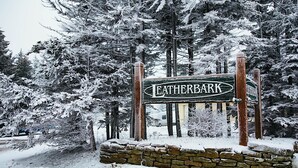 Un panneau recouvert de neige, portant l’inscription « LEATHERBARK » dans un style rustique, entouré de grands arbres à feuilles persistantes et d’un mur de pierres.