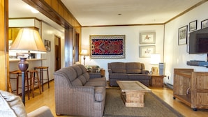A cozy living room with a sectional sofa, a coffee table, and a television. There are wooden cabinets and a rug on the floor. - Summit 304C, Snow White Summit 3 Br Walk to Slopes & Village (Snowshoe)
