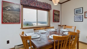 A dining area with a wooden table set for four, surrounded by wooden chairs. There is a large window with a view of mountains and a hanging light fixture above the table. - Summit 304C, Snow White Summit 3 Br Walk to Slopes & Village (Snowshoe)