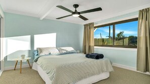 Ein Schlafzimmer mit einem Bett, Deckenventilator, einem Fenster mit Vorhängen und Blick ins Freie.