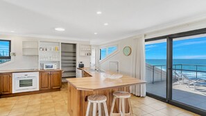 Une cuisine moderne dotée d’un îlot central, d’un coin repas et d’une grande porte-fenêtre coulissante donnant sur un balcon avec vue sur l’océan.