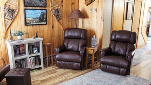 A room with wooden walls, two leather recliners, a small side table, a bookshelf, and a patterned rug.