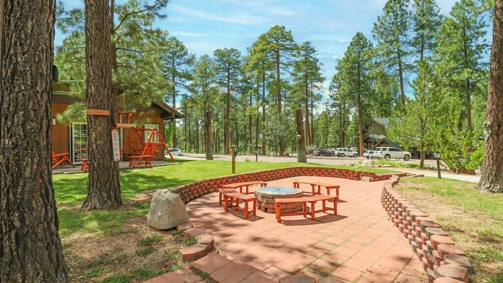 A patio area with a fire pit, benches, and a wooden table, surrounded by trees and a cabin.