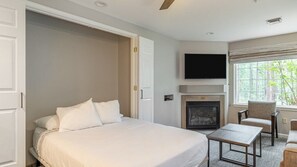 A bedroom with a bed, a TV mounted above a fireplace, and a window with a view of trees.