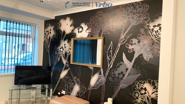 Une chambre moderne dotée d’une grande fenêtre, d’un téléviseur à écran plat, d’une table basse en verre, d’un banc rembourré et d’un mur décoratif orné de motifs floraux.