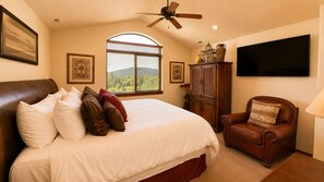 A bedroom with a large bed, a wooden cabinet, a leather armchair, a ceiling fan, and a TV mounted on the wall.