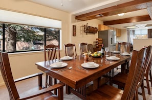 Une salle à manger avec une table en bois dressée pour quatre personnes, des chaises, une fenêtre donnant sur des arbres et, au fond, une cuisine.