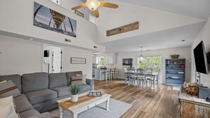 Un vaste salon avec un canapé gris, une table basse et un ventilateur de plafond. On aperçoit à l’arrière-plan un coin cuisine avec des meubles blancs et une table à manger.
