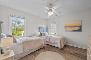 Une chambre avec deux lits, un ventilateur de plafond, de grandes fenêtres et un tableau représentant une plage.