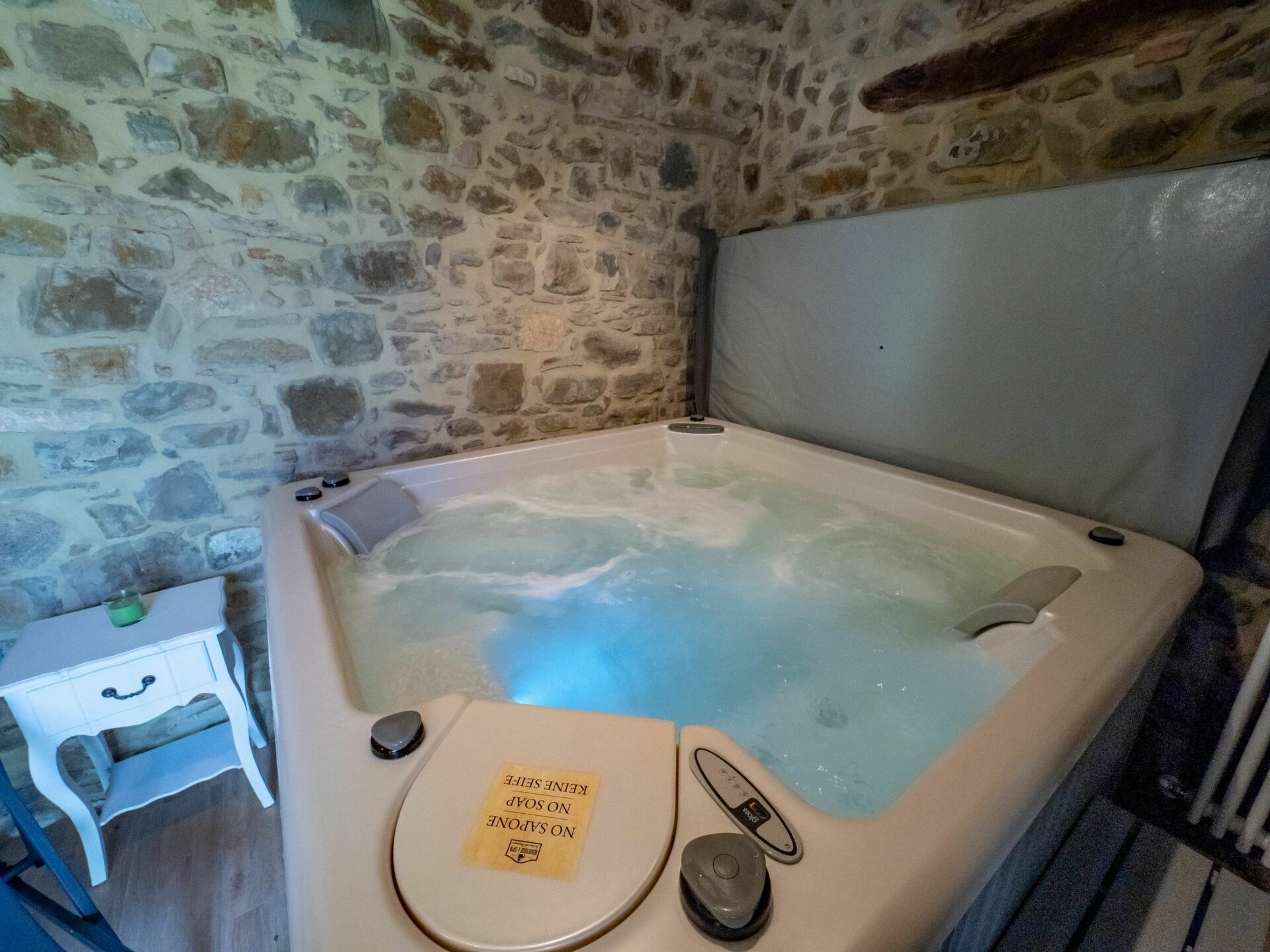 Une pièce aux murs de pierre, avec une baignoire remplie d’eau, une petite table de chevet sur laquelle se trouve une bouteille verte, et un panneau sur la baignoire.
