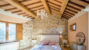 A bedroom with a stone wall, wooden beams, a chandelier, a bed with pillows, a dresser, and a mirror. - Stunning private villa for 4 people with hot tub, WIFI, A/C, TV, terrace and panoramic view (Seggiano)