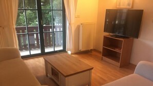 Una sala de estar con una mesa de centro de madera, dos sillones blancos, un televisor sobre un mueble bajo y un ventanal con cortinas.