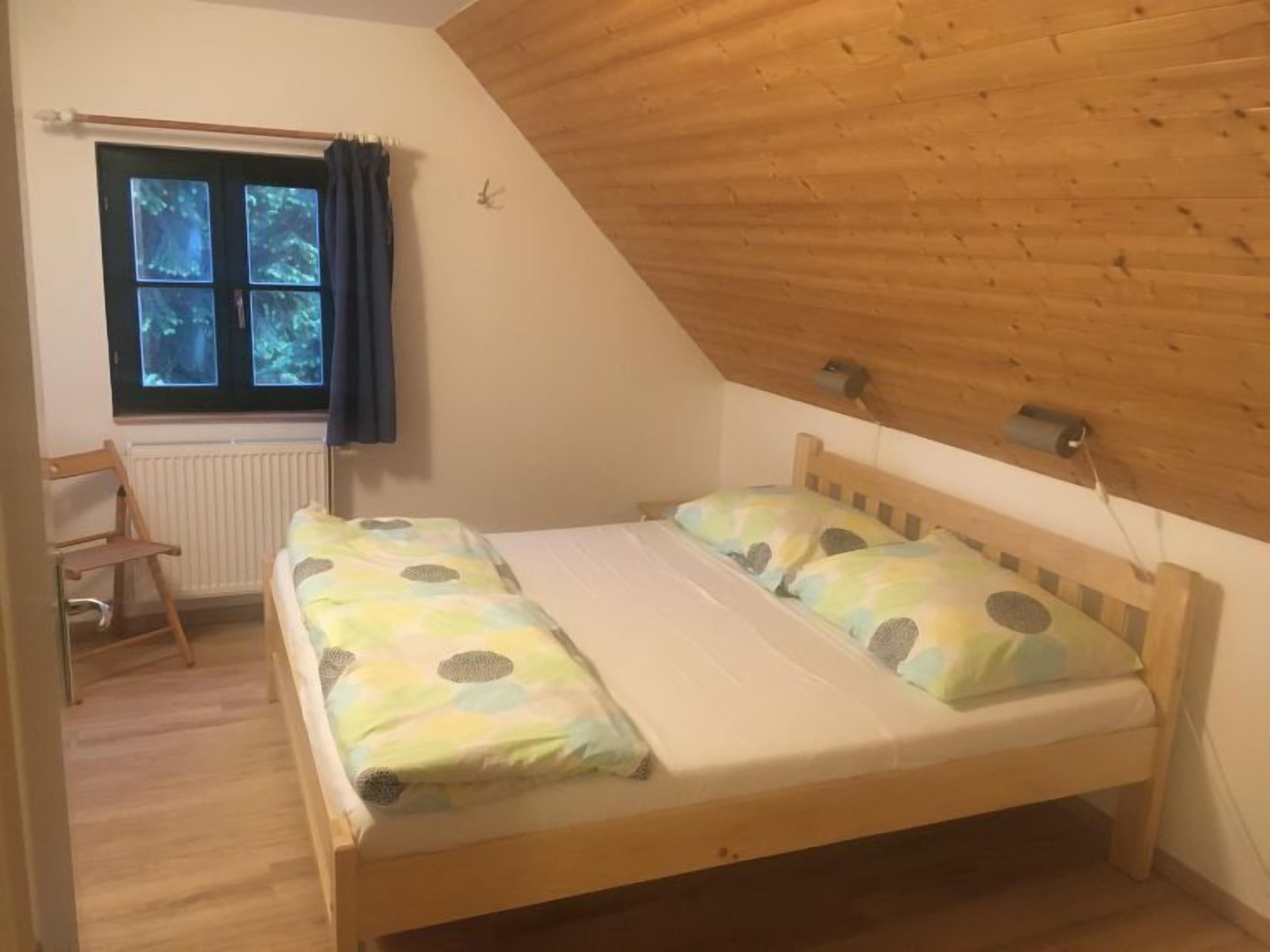 A small, single-bed room with a sloped wooden ceiling, a window with curtains, a radiator, and a wooden chair.