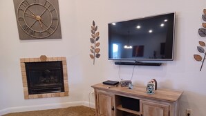 Una sala de estar con un televisor de pantalla plana montado en la pared, una chimenea con revestimiento de ladrillo y un mueble de madera con algunos objetos encima.