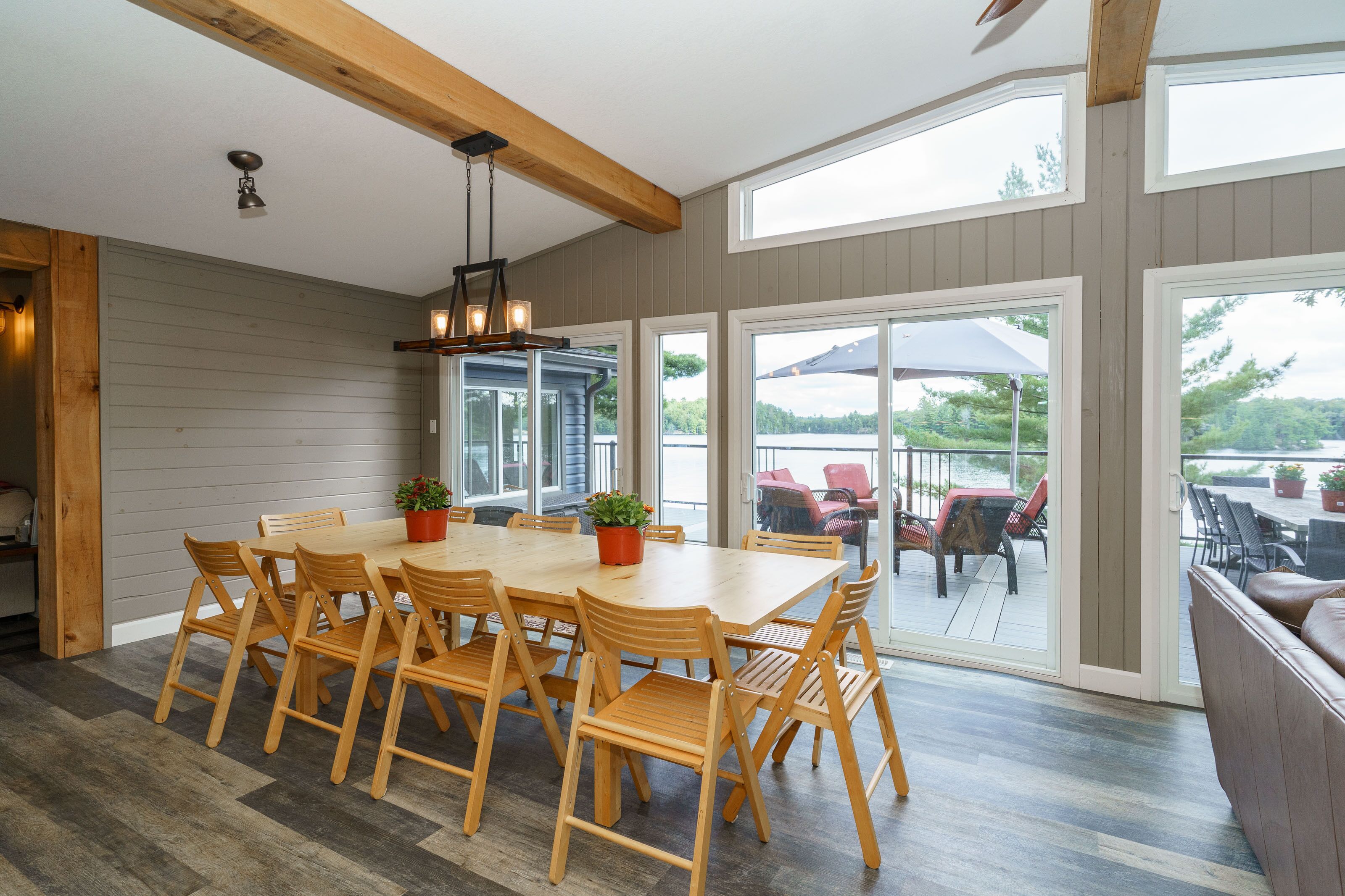 A dining area with a wooden table and chairs, a hanging light fixture, and large windows offering a view of the outdoors.