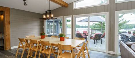 A dining area with a wooden table and chairs, a hanging light fixture, and large windows offering a view of the outdoors.