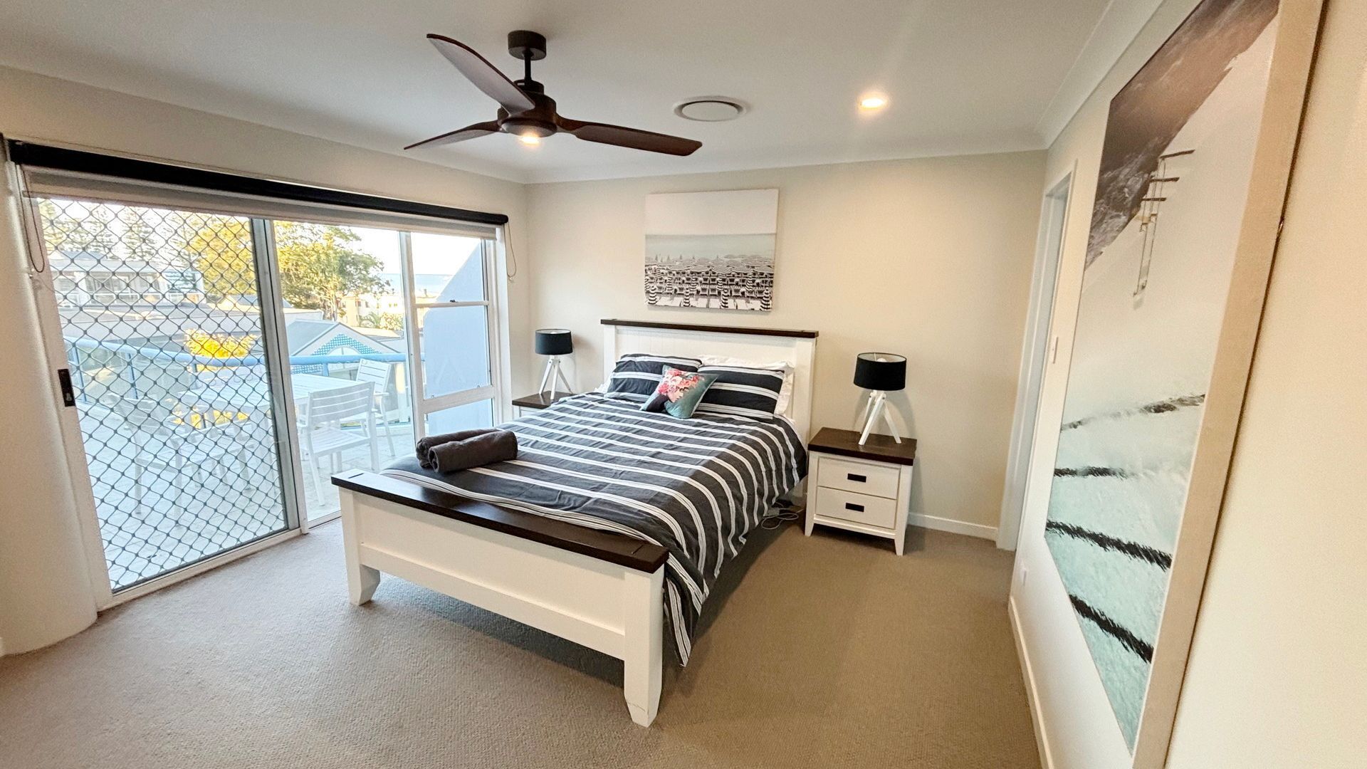 A bedroom with a bed, bedside tables, a ceiling fan, a window with a view, and a door.