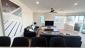 A modern living room with a flat-screen TV, a ceiling fan, and large windows. - Beach View Penthouse (SOUTH WEST ROCKS)