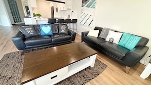 A modern living room with two black leather sofas, a wooden coffee table, and a kitchen in the background.