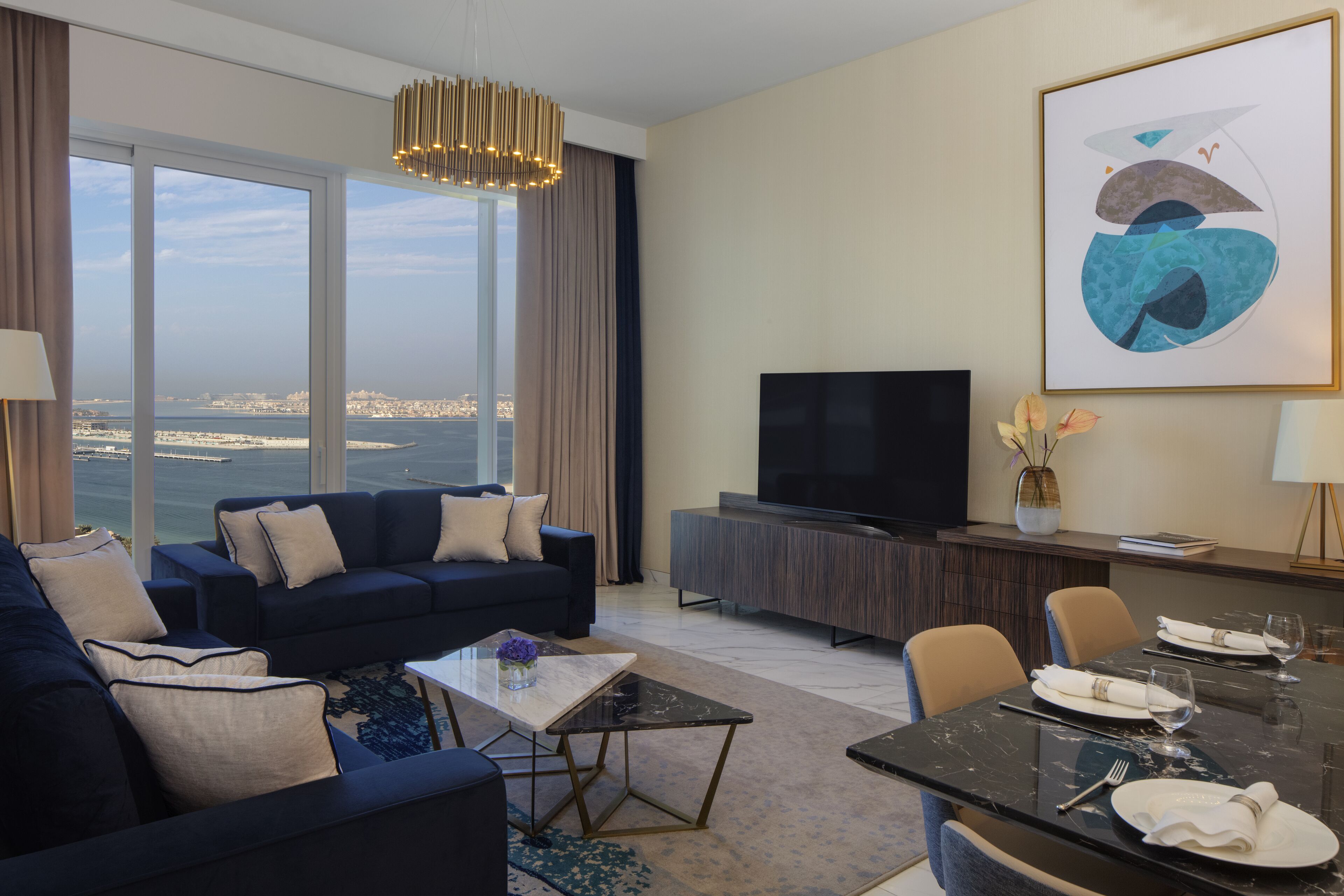 A modern hotel room with a large window offering a city view, a dark blue sofa, a flat-screen TV, a dining table set for two, and a minimalist decor.