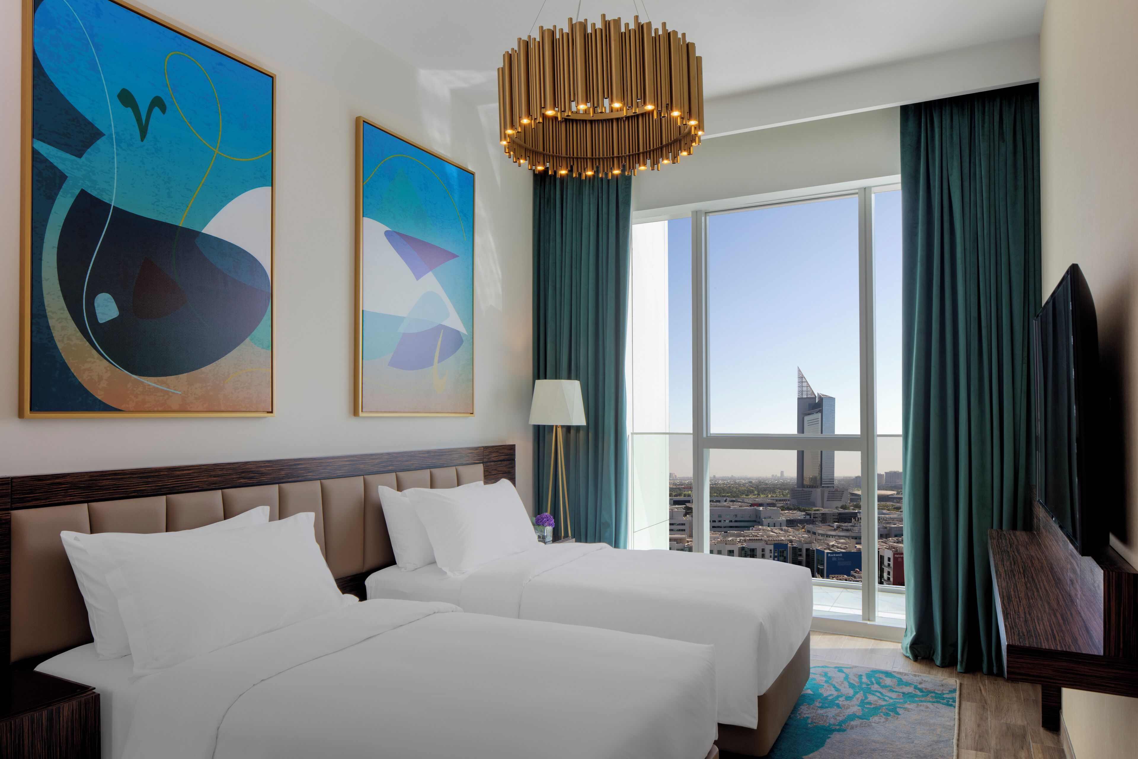 A hotel room with two beds, a large window with a city view, abstract paintings on the wall, and a modern chandelier.