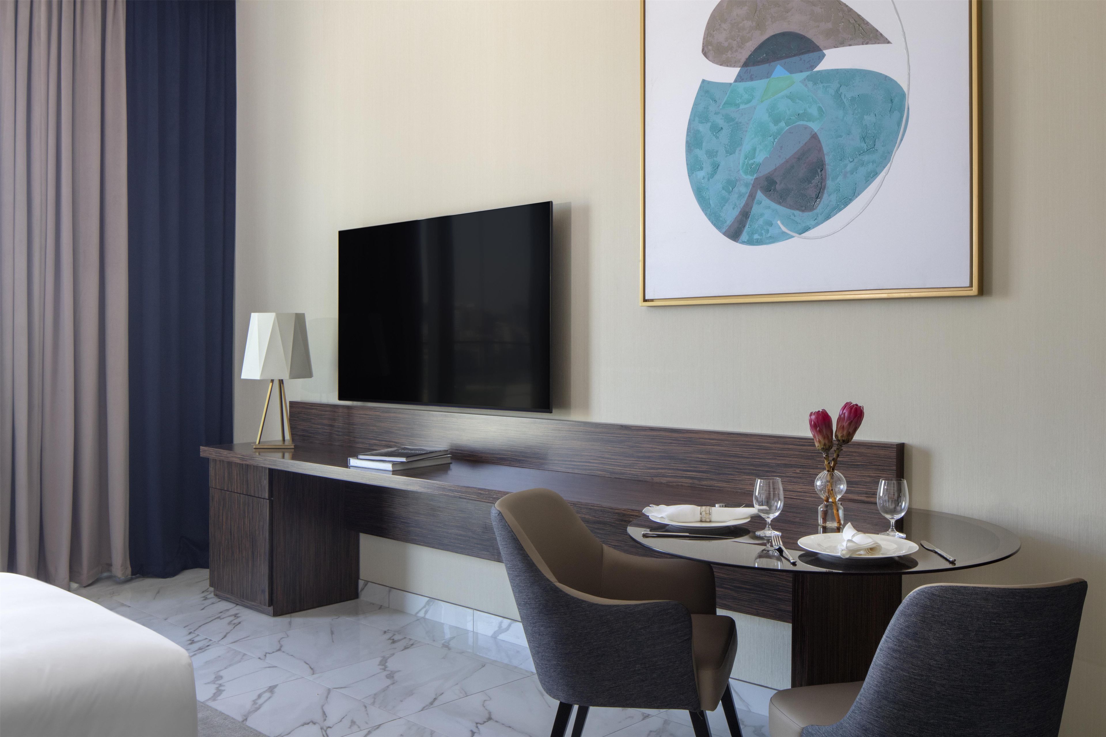 Habitación de hotel con televisor de pantalla plana en la pared, escritorio con lámpara, dos sillas, mesa de comedor con platos y vasos, y un cuadro abstracto grande.