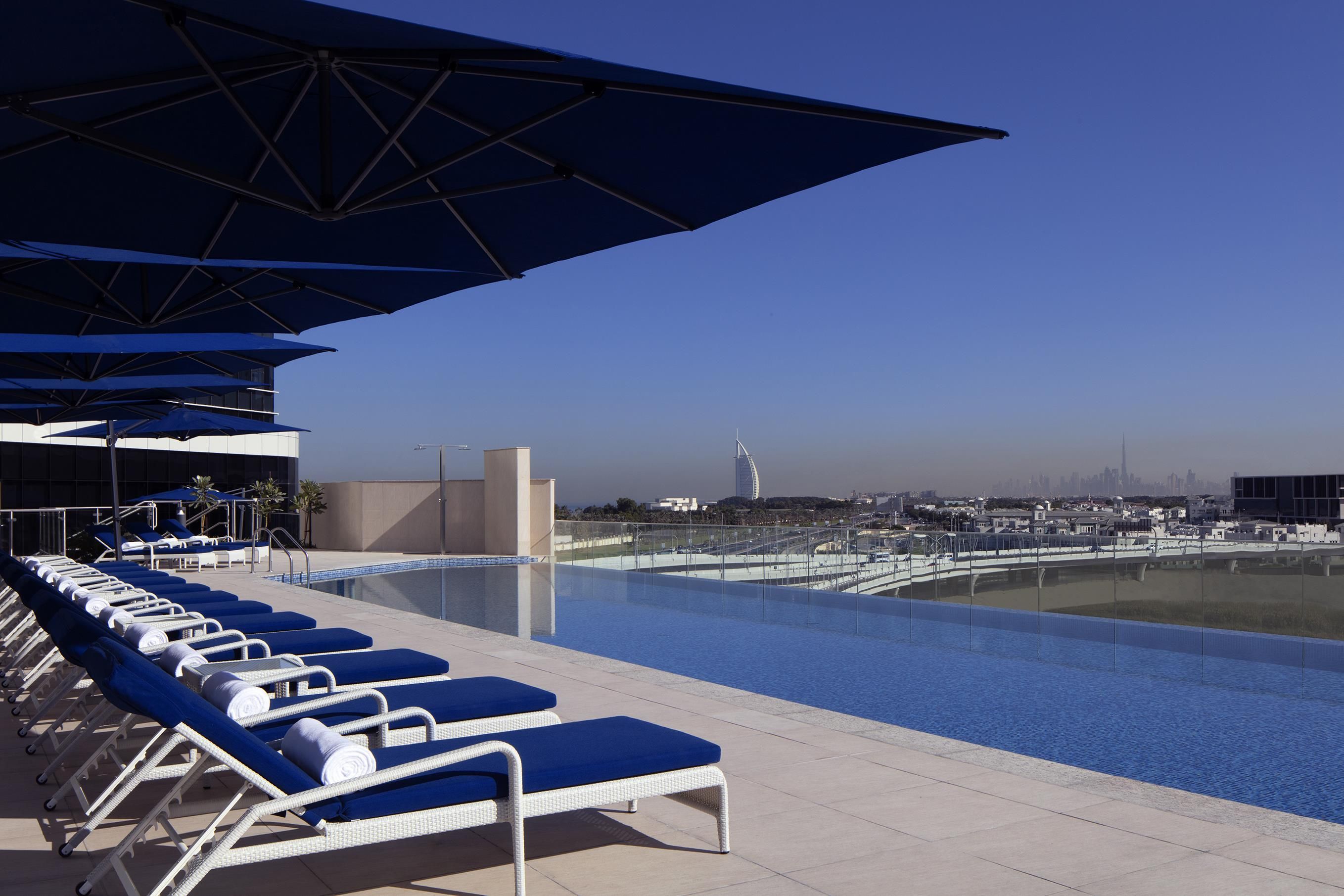 A poolside area with lounge chairs and umbrellas, a clear sky, and a distant cityscape.