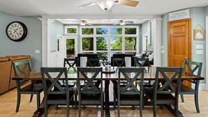 Un coin repas avec une table et des chaises en bois, un ventilateur de plafond et une vue sur l’extérieur grâce à de grandes fenêtres.