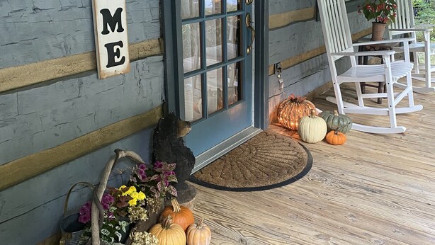 A wooden cabin with a 'WELCOME' sign, a front door, rocking chairs, and pumpkins on the porch.